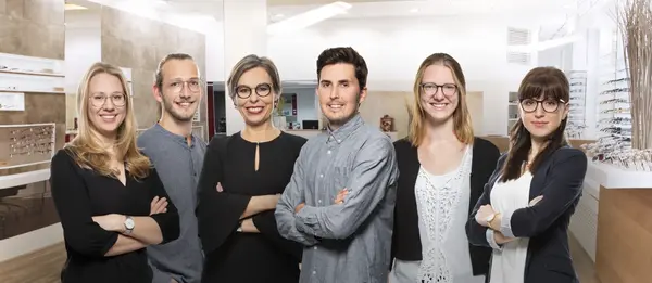 Sechs freundliche Optiker mit Brillen stehen im modernen Brillenfachgeschäft und lächeln.